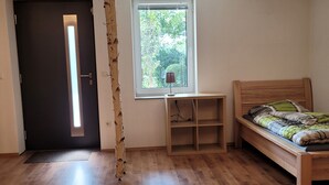 Quarto com piso de madeira, cama com cobertor verde e cinza, janela com vista para árvores, escrivaninha de madeira com abajur e porta com luminária.