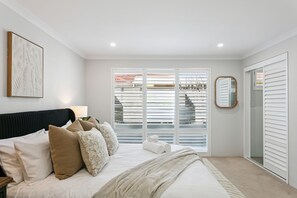 Ein Schlafzimmer mit Bett, Kissen, einer Nachttischlampe, einem gerahmten Kunstwerk, einem Fenster mit Jalousien und einem Spiegel.
