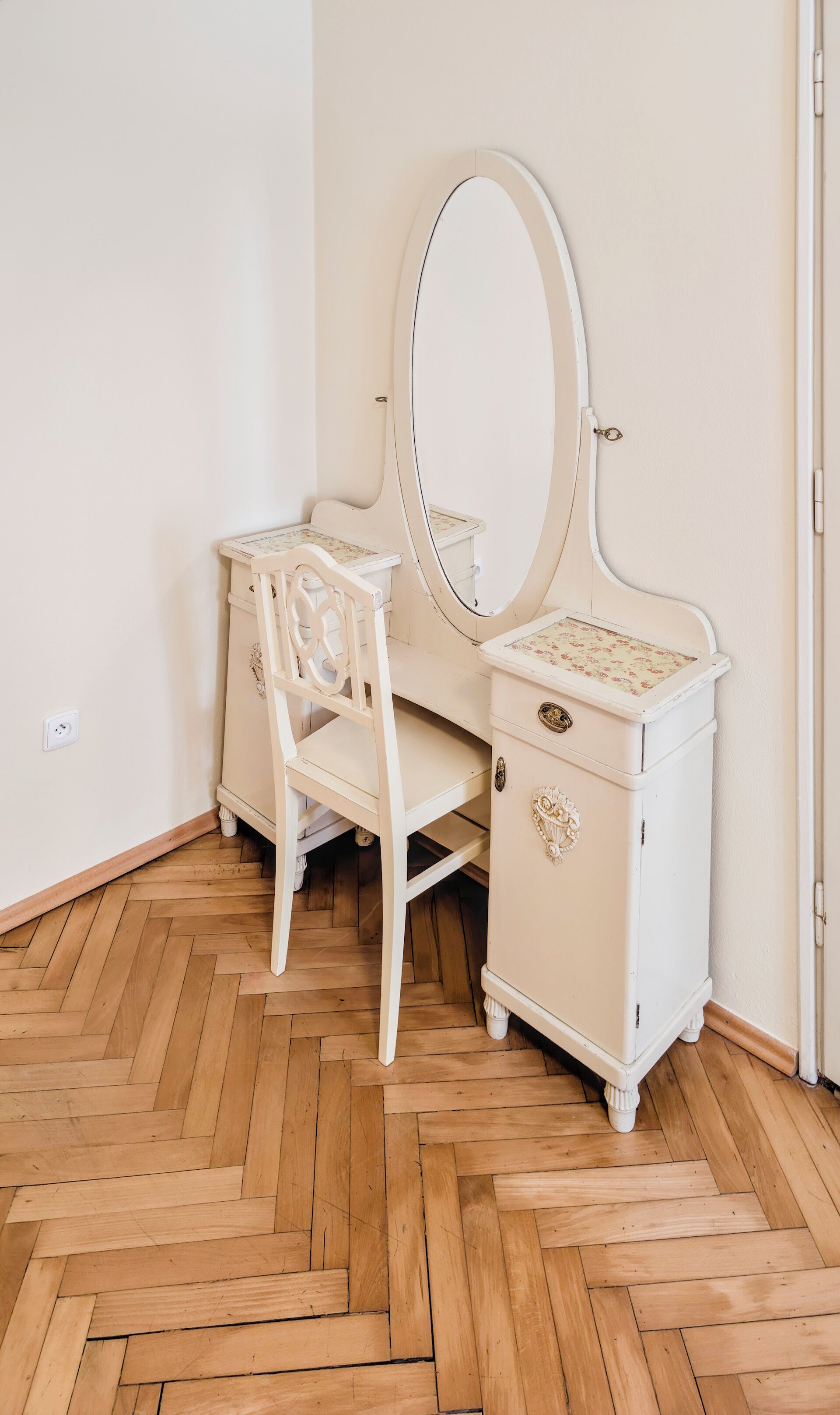 A white vanity with a mirror and a chair, wooden herringbone flooring, and a white wall.