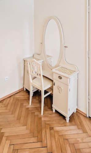 A white vanity with a mirror and a chair, wooden herringbone flooring, and a white wall.
