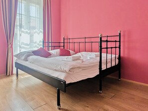 Una cama individual con estructura metálica y patas de madera, ubicada frente a una pared rosa con una ventana y cortinas.