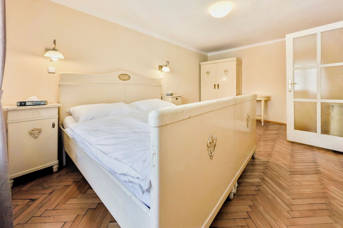 A single-bed room with a wooden herringbone floor, a bedside table with a lamp, a wardrobe, and a door with frosted glass panels.