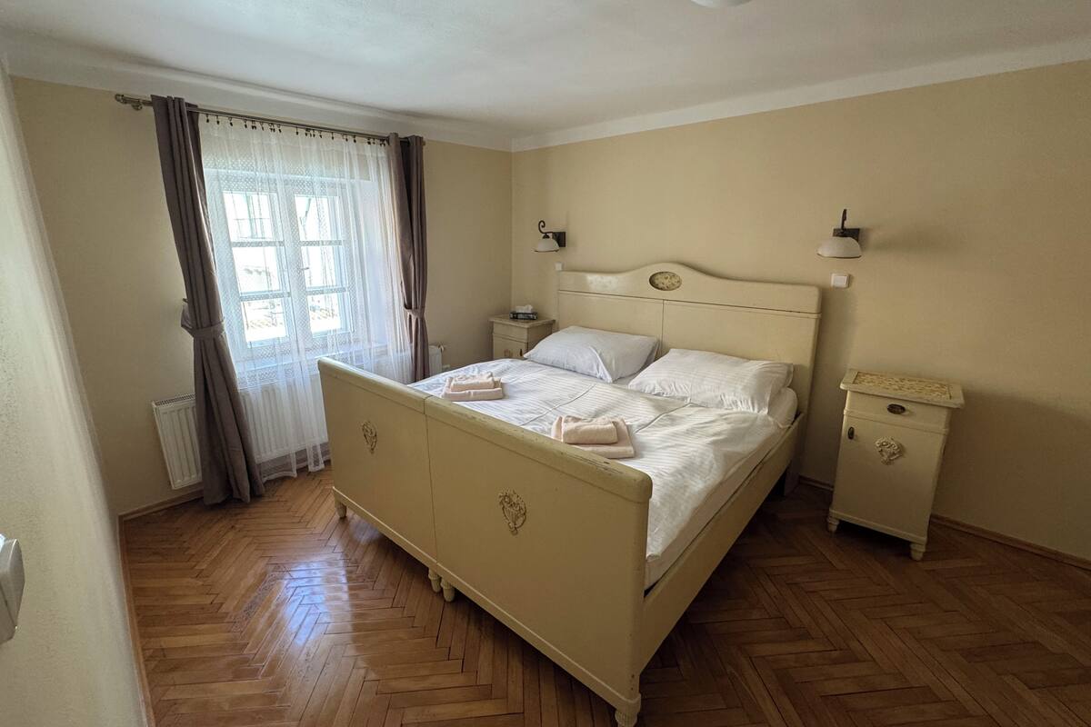 Un dormitorio con cama, mesita de noche, radiador, ventana con cortinas y suelo de madera.