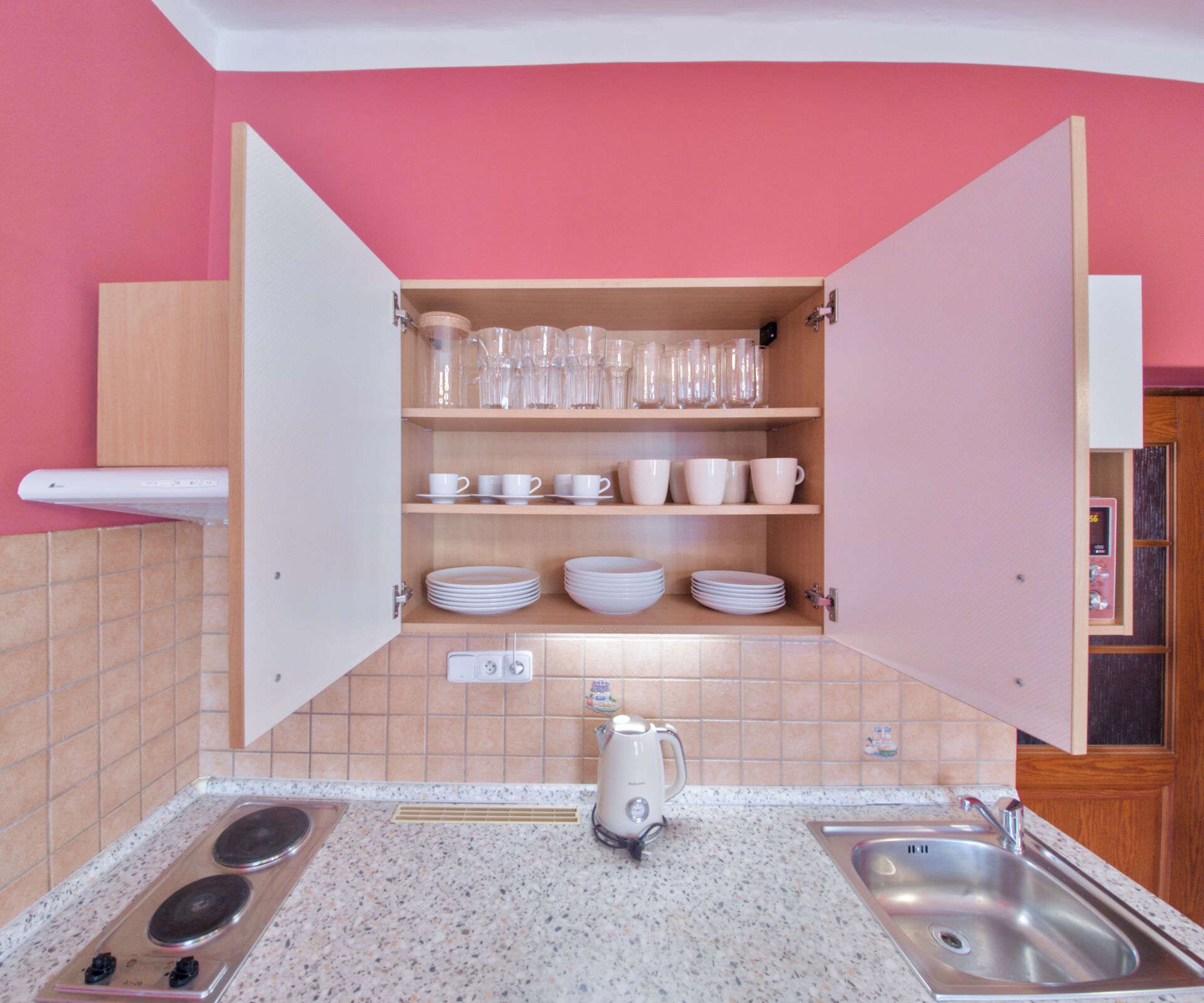 Cocina compacta con encimera, lavamanos y un armario con vajilla y cristalería.