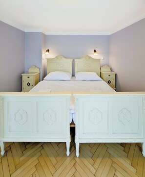 Ein Doppelbett mit weißem Kopfteil und Fußteil, flankiert von Nachttischen mit Lampen, in einem Zimmer mit Fischgrätparkett.