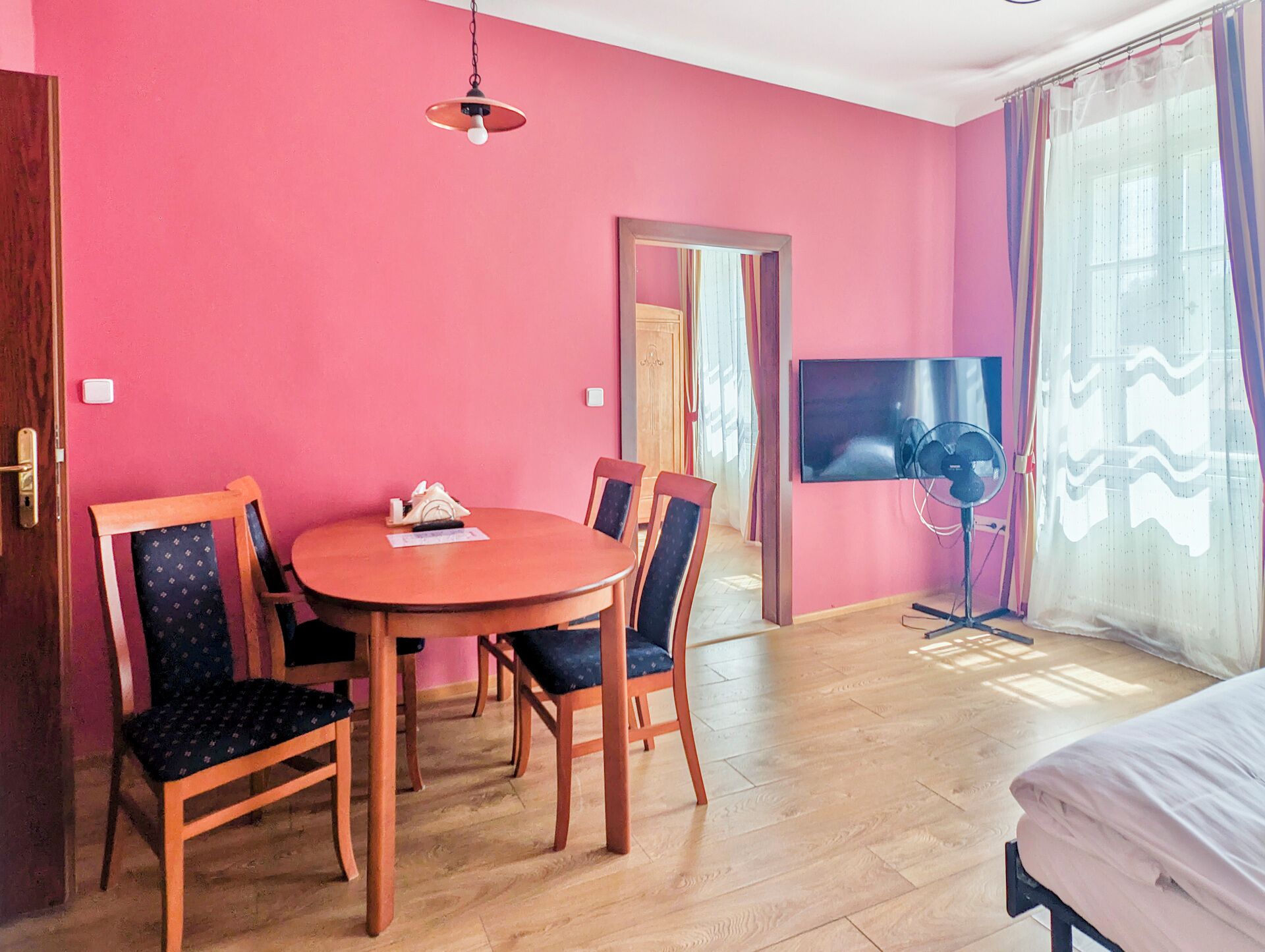 A room with a round wooden table, four chairs, a television on a stand, and a bed.