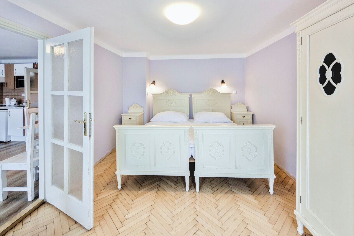 A double bed with a white headboard and two bedside tables, a wooden herringbone floor, and a white door with a decorative glass panel.