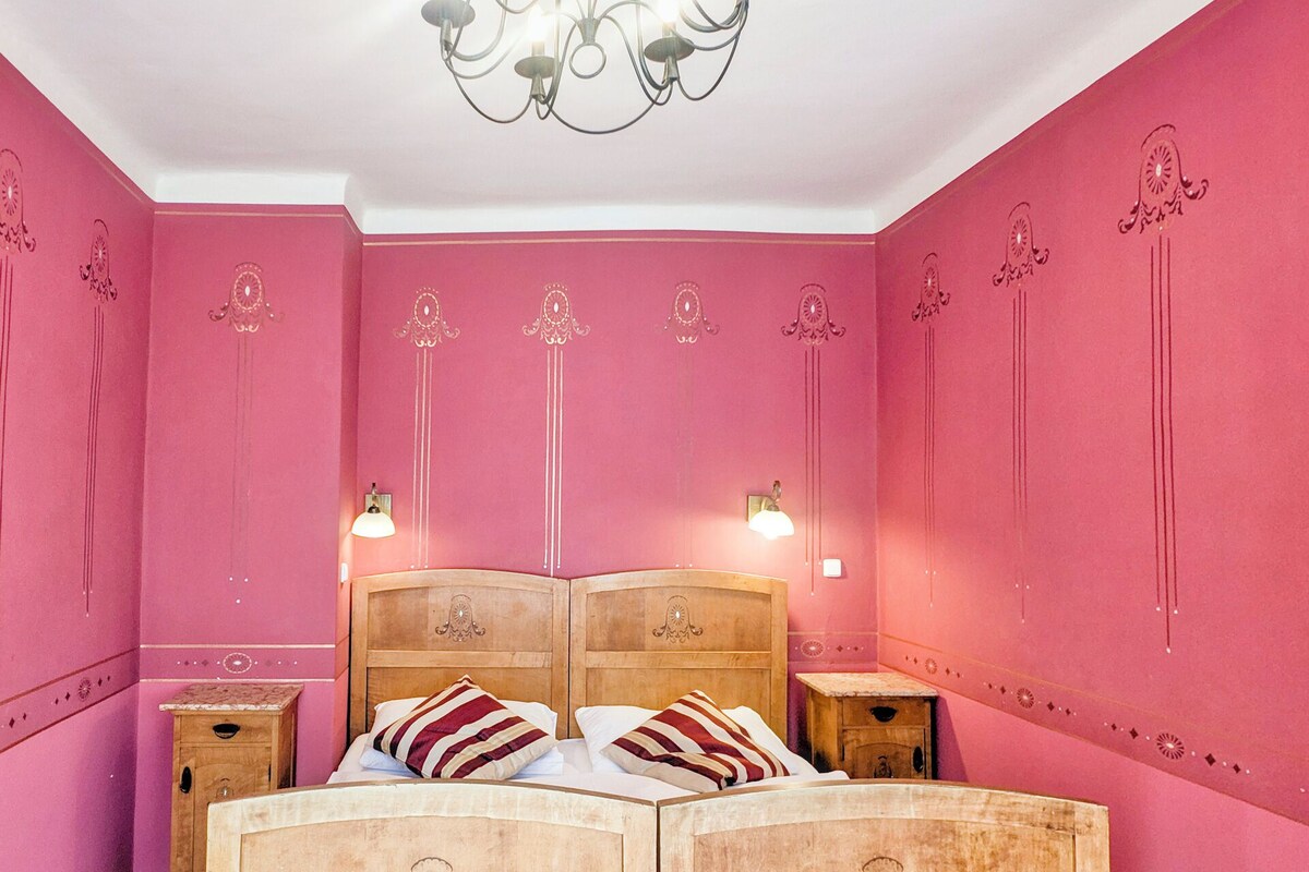 A room with a bed, two bedside tables, a chandelier, and pink walls.