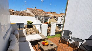 En udendørs terrasse med en sofa, stole, et bord med frugt og en kop, samt udsigt til bygninger i baggrunden.