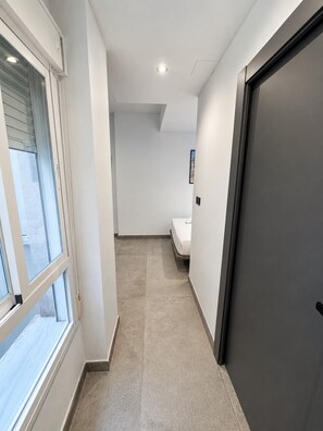 Ein schmales Gang mit einem Fenster links, einem Bett im Hintergrund und einer Tür rechts.
