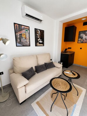 Ein modernes Wohnzimmer mit einem beige Coloured Sofa, zwei gerahmten Bildern, einer Stehlampe und einer kleinen Küchenzeile.
