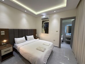 A modern hotel room with a large bed, bedside tables, an air conditioning unit, a painting, and a bathroom visible through an open door. - 201 JRE Apartment With Stunning Boulevar (Amman)