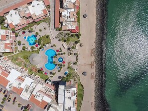 Luftaufnahme eines Küstenresorts mit mehreren Schwimmbecken, einem Strand und angrenzenden Gebäuden.