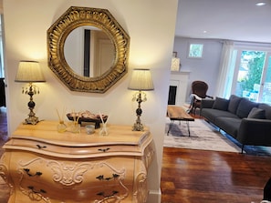 Een kamer met een grote, versierde spiegel, twee lampen, een houten dressoir, een bank en een open haard.