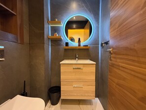 Et moderne badeværelse med en trækommode, et rundt spejl med blå lys og en hylde med toiletartikler.