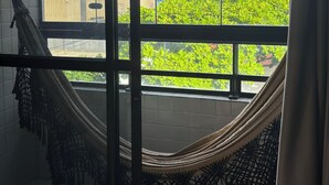 Une pièce dotée d’une grande fenêtre, d’un hamac et offrant une vue sur des bâtiments et de la verdure.