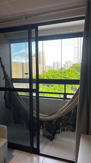 A room with a large window, a hammock, and a view of buildings and greenery. - Bellenus - balcony, hammock, comfort. (João Pessoa)