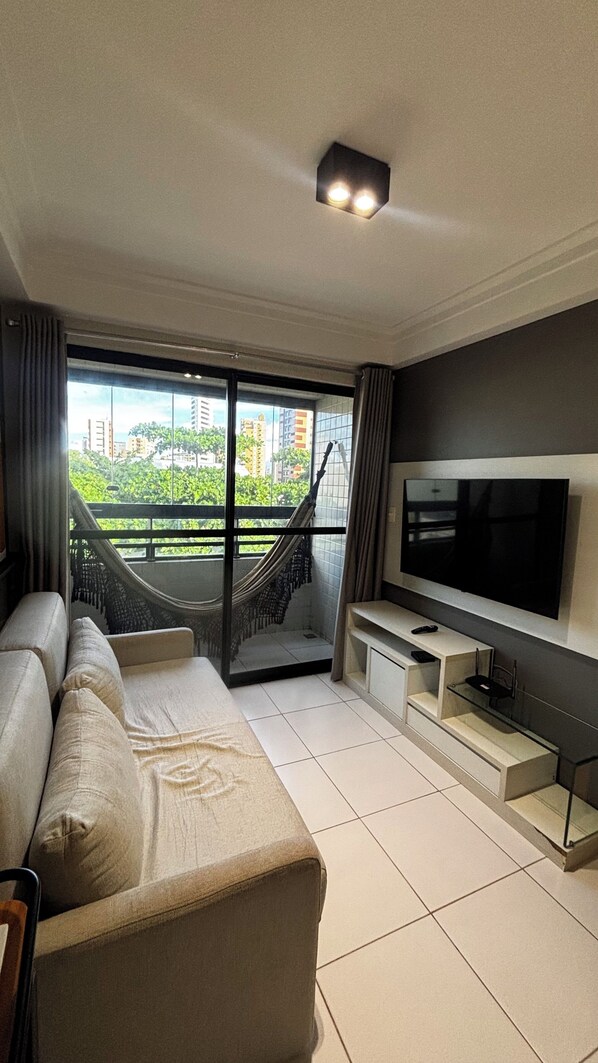 A modern living room with a sofa, TV, and a view of greenery and buildings. - Bellenus - balcony, hammock, comfort. (João Pessoa)