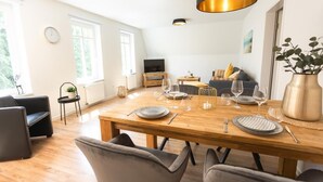 A dining area with a wooden table set for four, a television in the background, and a sofa with cushions. - Helga's Stadthaus (Nordhorn)