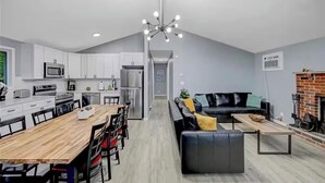 A modern kitchen with white cabinets, a dining area with a wooden table, and a living room with a black sofa and a fireplace. - Poconos Chalet, Game Room, Hot Tub & Firepit (Tobyhanna)