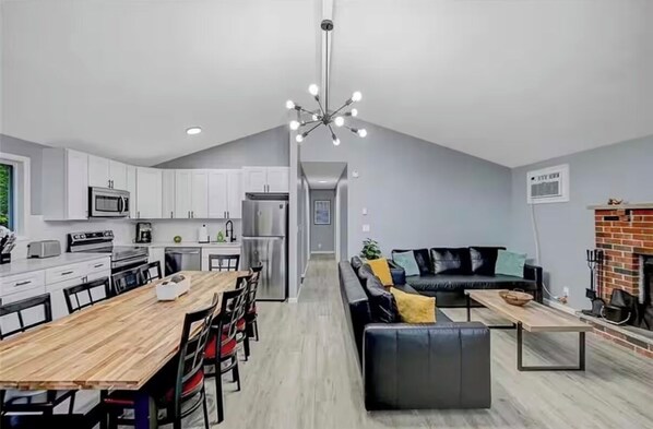 Een moderne keuken met witte kastjes, een eethoek met een houten tafel, en een woonkamer met een zwarte bank en een open haard.