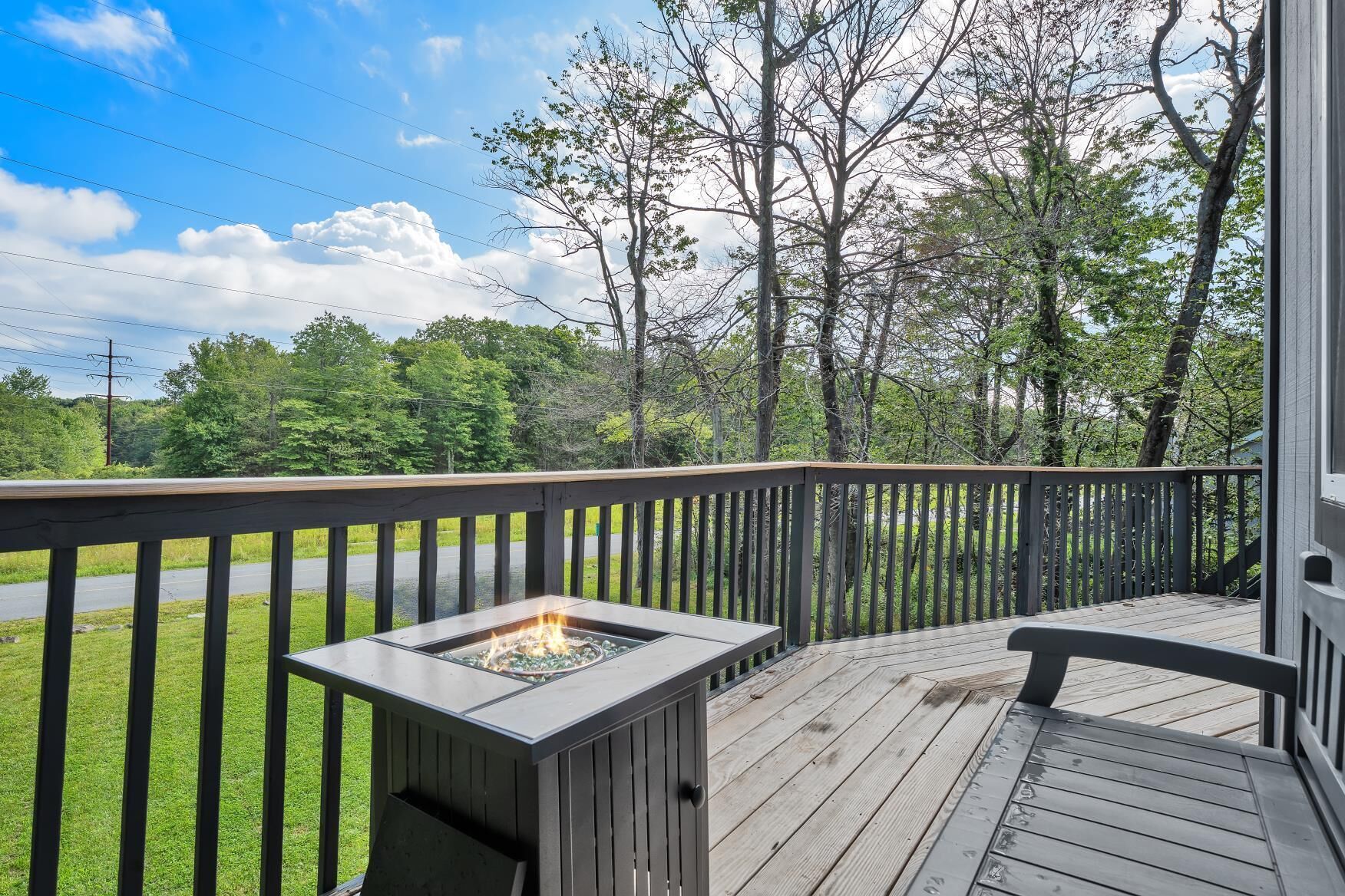 A wooden deck with a fire pit, surrounded by trees and a road.
