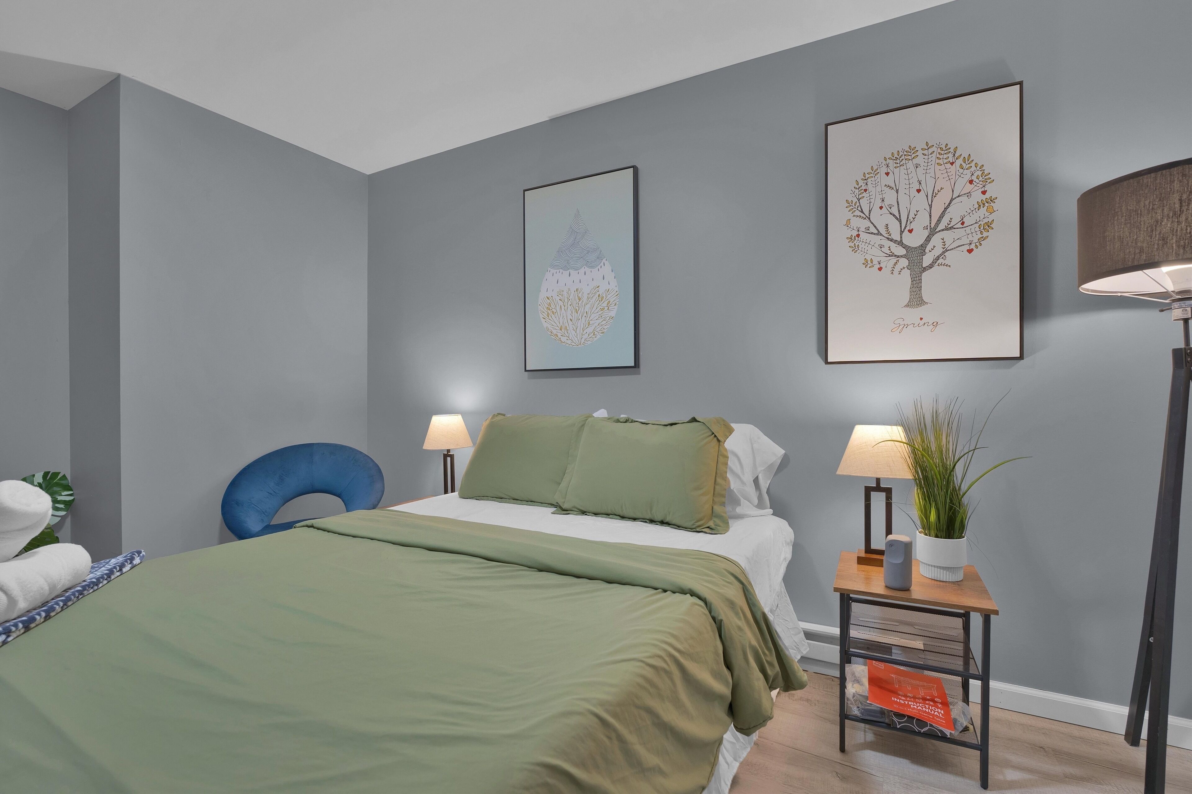 Ein Schlafzimmer mit einem Bett, zwei Wandlampen, einem blauen Sessel, einer Topfpflanze und zwei gerahmten Kunstwerken.