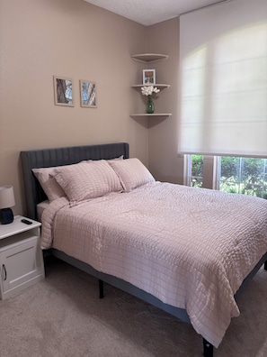 A bedroom with a bed, bedside table, lamp, wall art, and a window with blinds.