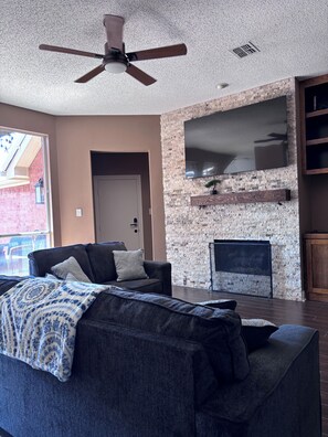 A living room with a stone wall, a fireplace, a ceiling fan, and a large TV. - Private Garden Retreat in Frisco TX - 3 bedroom and 2 full baths (Frisco)