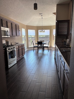 A kitchen with dark wood flooring, a dining area with a table and chairs, and a view through large windows.
