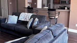 Un salon avec un canapé et des fauteuils, un coin cuisine équipé d’appareils modernes, et une porte donnant sur une autre pièce.