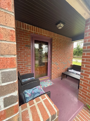Un bâtiment en briques avec une porte violette, deux chaises en osier et un banc sur un porche abrité.