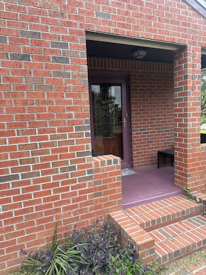 Un bâtiment en briques avec une porte violette, un petit banc et des marches menant à un porche.