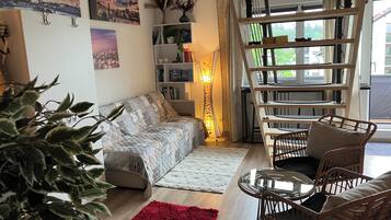 A living room with a sofa, a red rug, a glass table, wicker chairs, a bookshelf, and a window with a view.