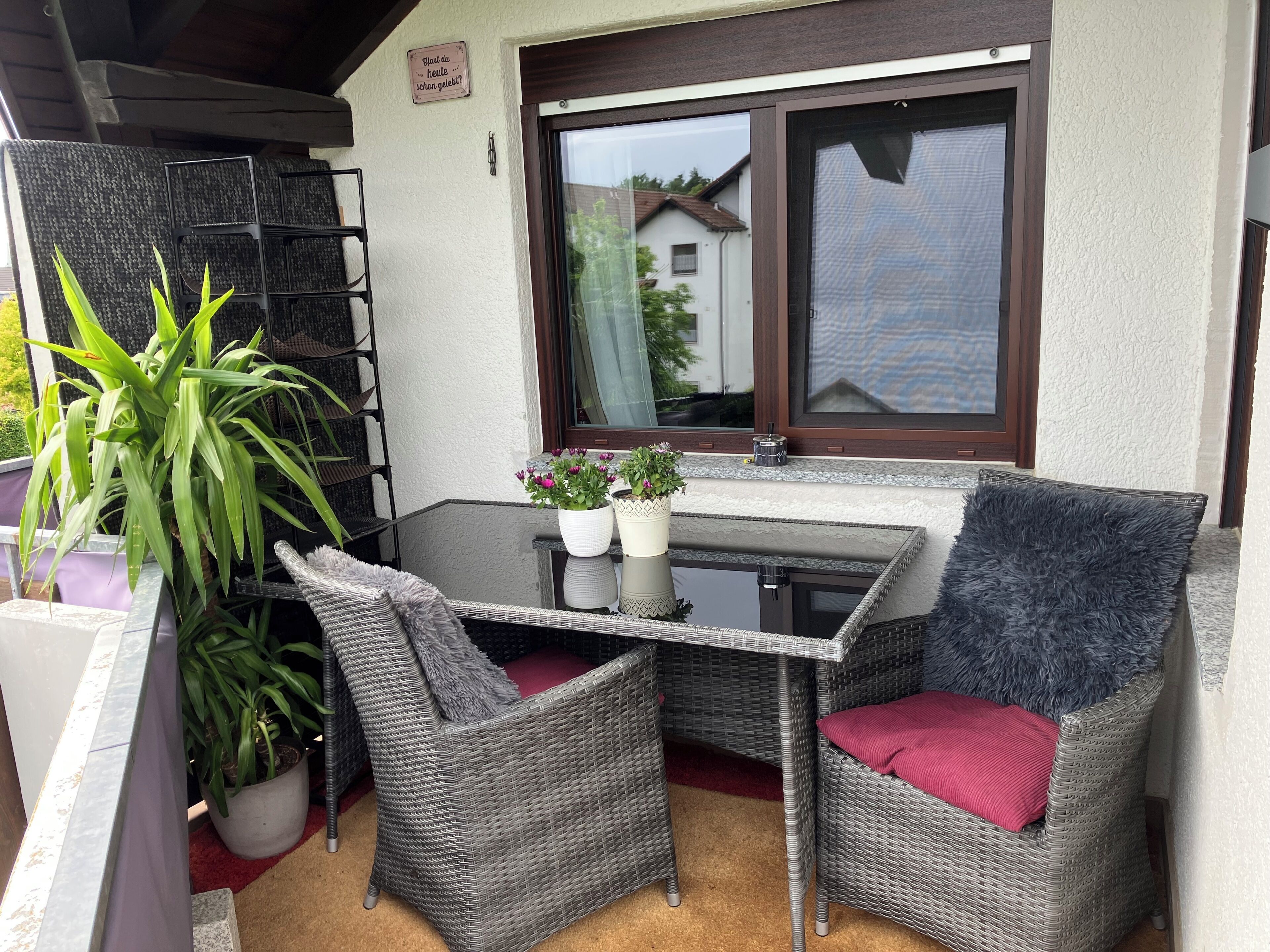 A balcony with wicker furniture, potted plants, and a window with a view of houses.
