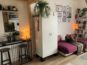 Ein kleines Zimmer mit einem Bett, einem Schreibtisch mit Hocker, einem Kleiderschrank, einem Spiegel und einer Wand mit gerahmten Bildern sowie einem Regal mit Büchern und Pflanzen.