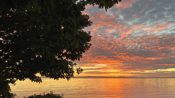 Pôr do sol sobre águas calmas, com terreno rochoso e vegetação densa.