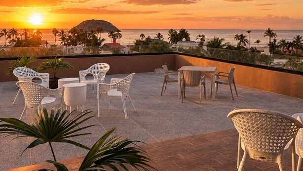 A rooftop terrace with outdoor seating, a view of the sunset over the ocean, and palm trees. - Boutique apartment with ocean view, pool, 2 blocks from the beach! (Rincón de Guayabitos)
