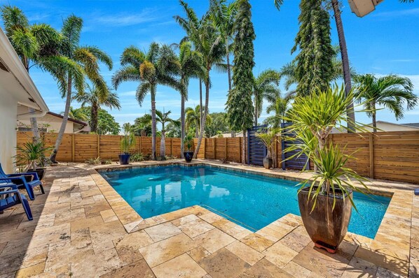 A swimming pool with a wooden deck, surrounded by palm trees and a wooden fence.