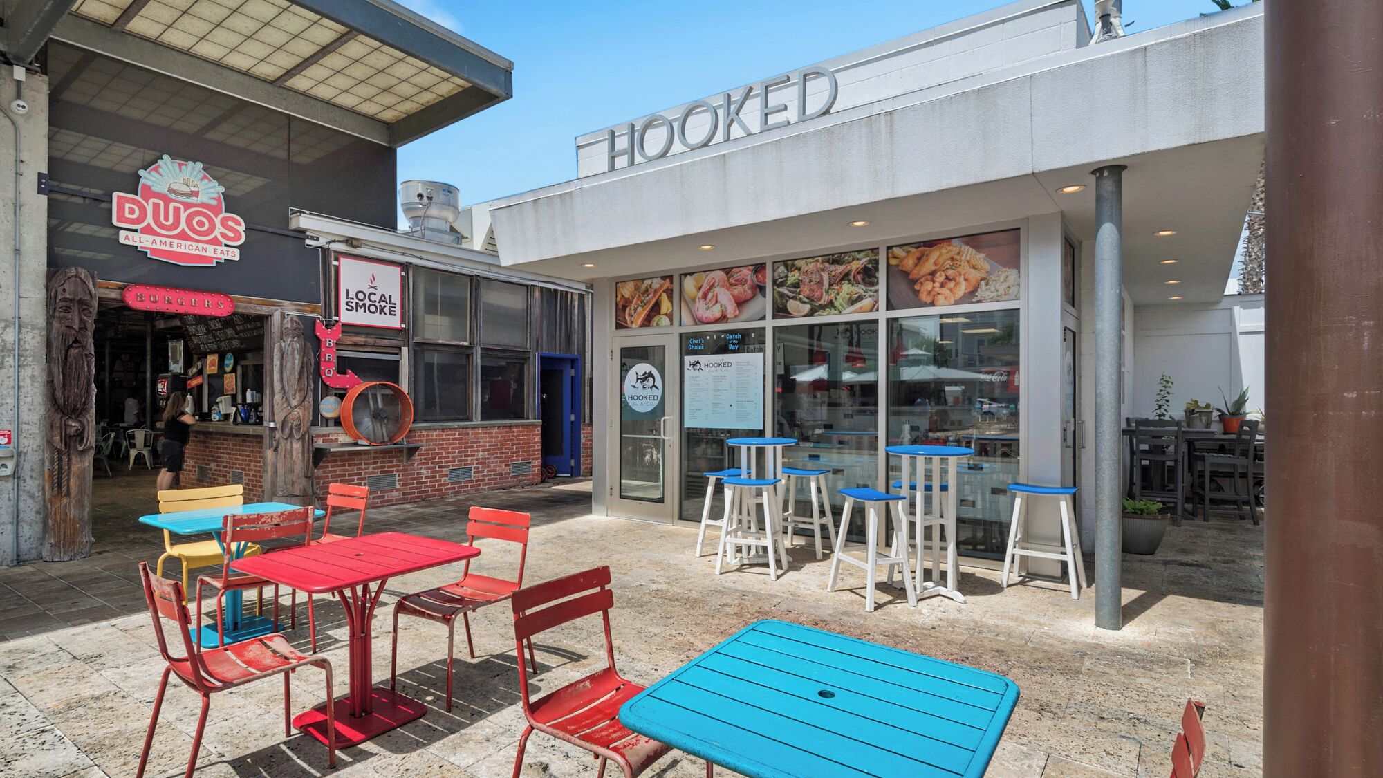 An outdoor seating area with tables and chairs, a sign for 'HOOKED' restaurant, and a 'DUOS ALL-AMERICAN EATS' sign.