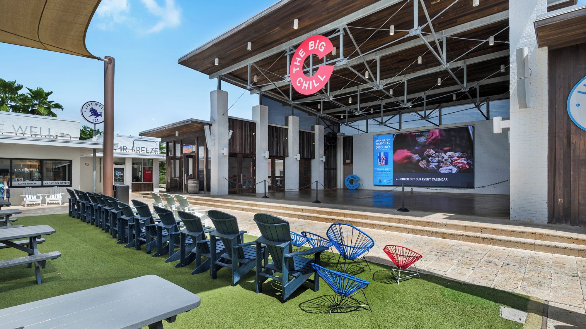 Un espacio al aire libre con sillas y mesas, un escenario con una pantalla grande y un cartel que dice "The Big Chill".