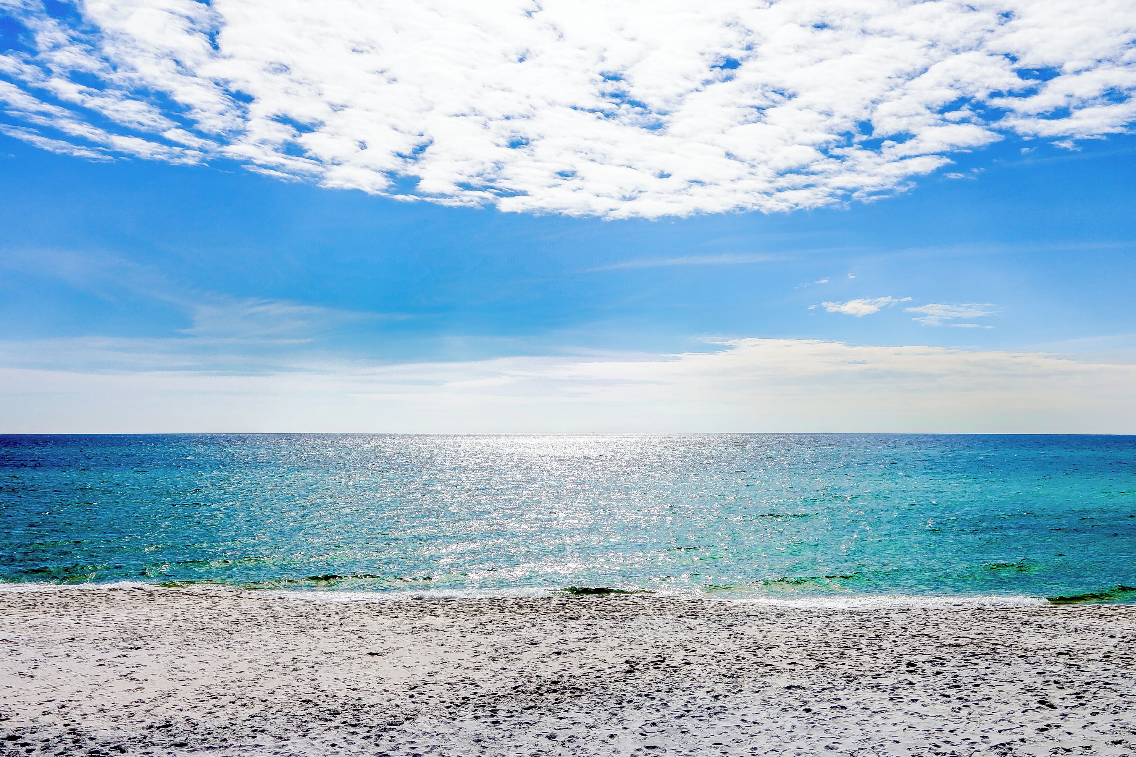 Une plage à l’eau d’un bleu limpide et au rivage sablonneux, sous un ciel partiellement nuageux.