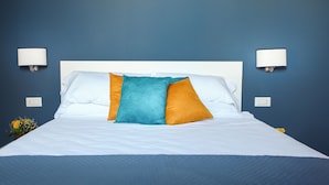 Una cama bien hecha con almohadas blancas y anaranjadas, una almohada de acento azul y dos lámparas fijadas a la pared.