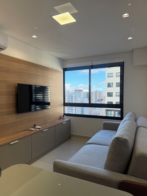 A modern living room with a flat-screen TV, a sofa, a wooden cabinet, and a large window offering a city view. - Modern studio apartment in the city center with air conditioning, garage, swimming pool, and gym. (Curitiba)