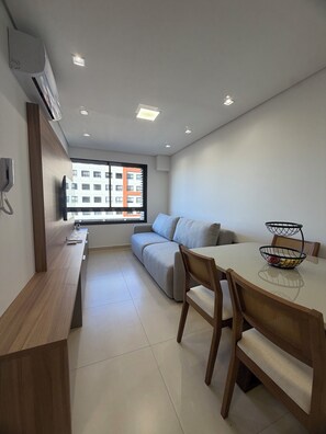 A modern, compact living space with a sofa, dining table, and chairs, a built-in TV, and a window with a city view. - Modern studio apartment in the city center with air conditioning, garage, swimming pool, and gym. (Curitiba)