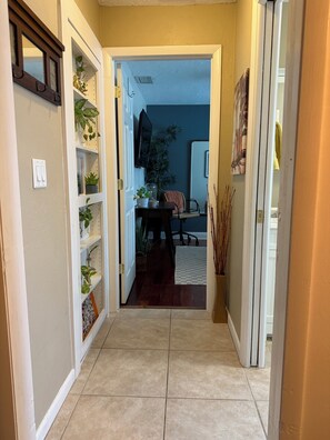 Un pasillo con una puerta que da a una habitación, un televisor montado en la pared y una estantería con plantas.