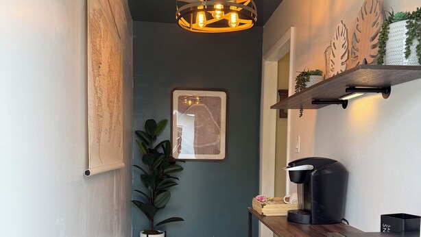 A narrow hallway with a checkered rug, a hanging light fixture, a potted plant, a mirror, a wooden shelf with plants, a coffee maker, and a microwave. - Comfort in the Cape (Cape Coral)