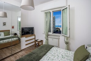 Ein Schlafzimmer mit einem Bett, einem Fernseher, einem Fenster mit Vorhängen, einem Heizkörper und einem kleinen Tisch mit Büchern.
