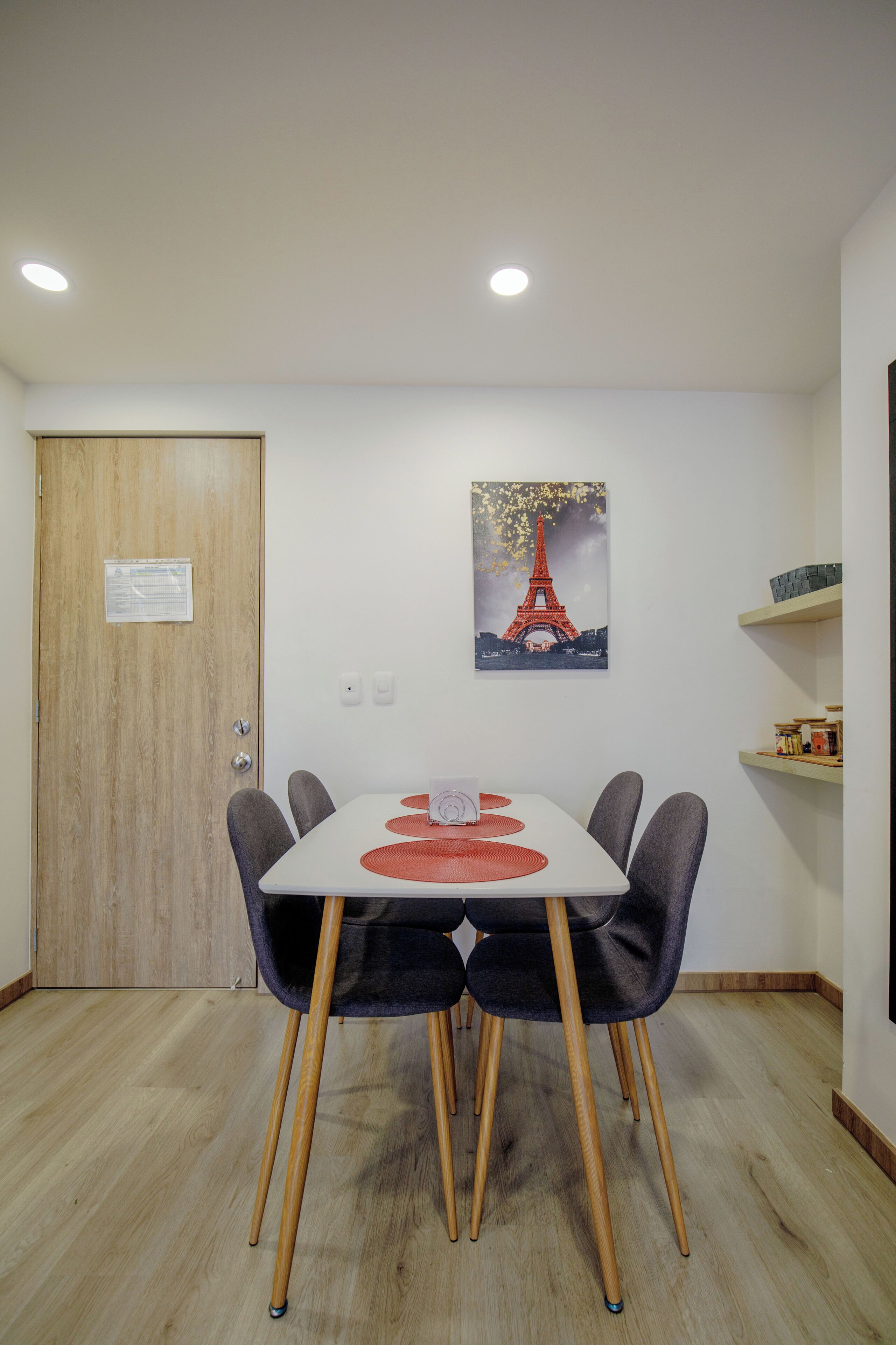 A dining area with a table, chairs, a wooden door, a wall-mounted picture of the Eiffel Tower, and a shelf with items.