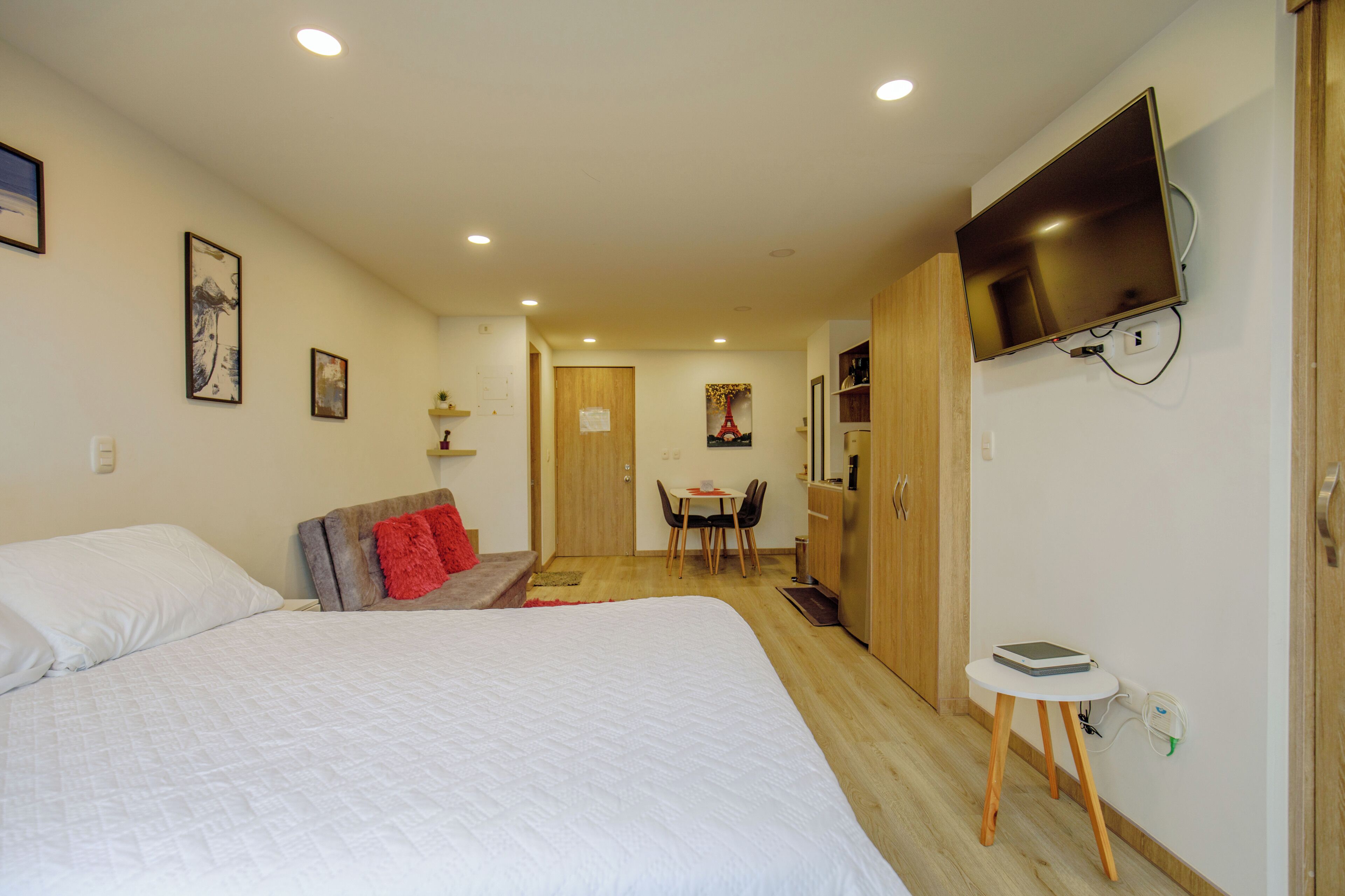 A hotel room with a bed, a TV mounted on the wall, a desk with a chair, and a small table with a power outlet.